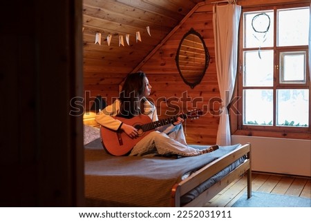 Creative millennial girl sitting on bed holding guitar in hands and looking out of window, waiting for inspiration. Musical Instrument and mental health. Hobby and recreational activity concept