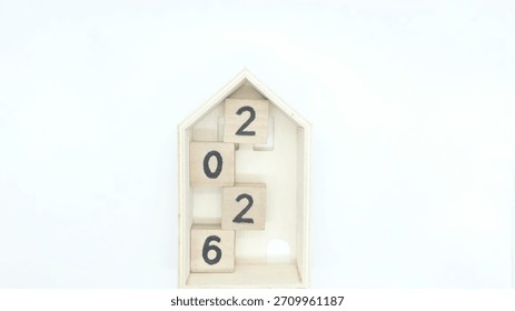 A creative and minimalist composition featuring four wooden blocks arranged vertically inside a small house-shaped wooden frame, forming the number 2026.