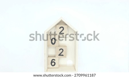 A creative and minimalist composition featuring four wooden blocks arranged vertically inside a small house-shaped wooden frame, forming the number 2026.