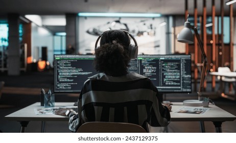 Creative Office: Professional Black Female Programmer Uses Headphones, Working on Desktop Computer. Focused Coder, Software Engineer Developing App, Video Game. Listening to Podcast, Music. Back View.