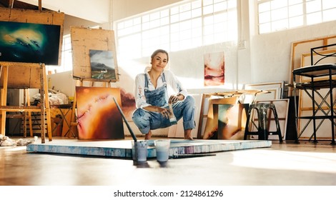 Creative painter squatting close to her painting in an art studio. Artistic young woman working on a new painting on the floor. Cheerful young artist looking at the camera while holding a paintbrush.