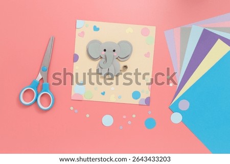 Creative paper craft featuring cheerful gray elephant with pink cheeks on yellow sheet, surrounded by colorful cut paper. Great for kids activities, craft blogs, DIY tutorials, or classroom materials.