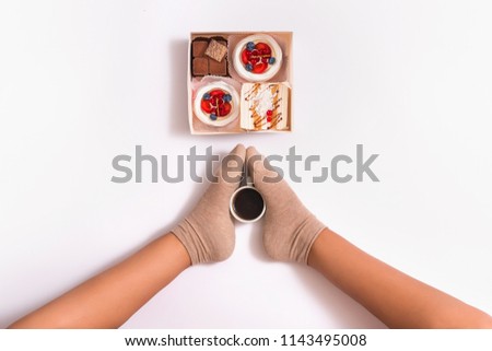 
Creative photo of box with cakes and cup of coffee between female feets on white background. Top view
