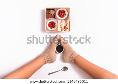 
Creative photo of box with cakes and cup of coffee between female feets on white background. Top view