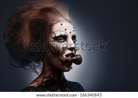 A creative photo of a queens painted pierced face holding a chess king in her mouth.