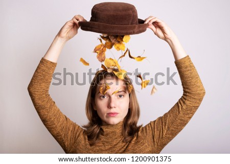 Creative portrait of a beautiful woman with a hat full of autumn leaves. She is empting leaves on her head.