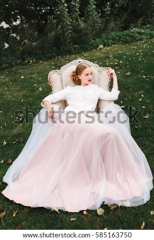 creative portrait of woman in a beautiful long pink dress sits on the couch which is on the grass on the background of a summer garden, fashion hairstyle and makeup, plein air photoshoot