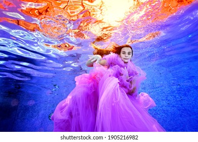 A Creative Portrait Of A Young Girl In A Luxurious Pink Dress, Who Swims And Plays Underwater In The Pool Against The Background Of Bright Light On The Surface Of The Water. Fashion Photography.