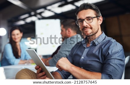 The creative process. Portrait of a man sitting at a table in an office using a digital tablet with colleagues working in the background.