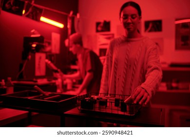 Creative process of two young photographers working with analog film while developing photo prints in darkroom lit by red lights copy space