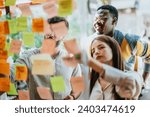 Creative professionals work on a project, utilizing a glass board and sticky notes. Successful teamwork and communication lead to a modern, persistent corporation.