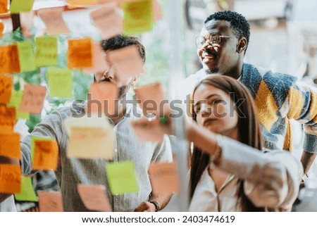 Creative professionals work on a project, utilizing a glass board and sticky notes. Successful teamwork and communication lead to a modern, persistent corporation.