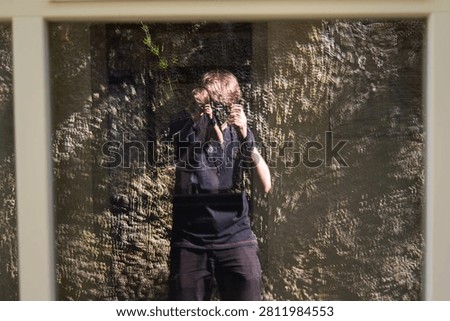 A creative self-portrait reflection in a glass windowpane, showing a photographer holding a camera, visually superimposed over the rugged texture of a sunlit stone wall.