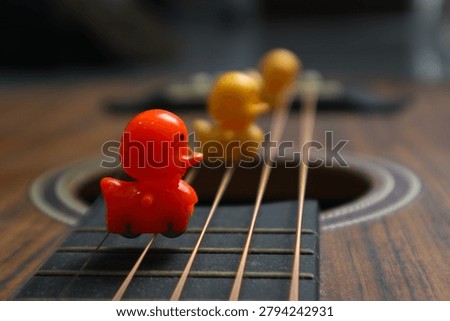 A creative shot featuring a bright orange plastic duck followed by yellow ducks, balanced on the strings of an acoustic wooden guitar. This image represents themes of leadership, standing out from the crowd, and musical inspiration. The shallow depth of field emphasizes the front duck against a blurred background.