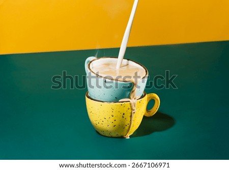 Creative shot of hot coffee in stacked colourful ceramic mugs with milk being poured in. Overflowing coffee with steam on green and yellow background.Morning energy, caffeine, and creative photography