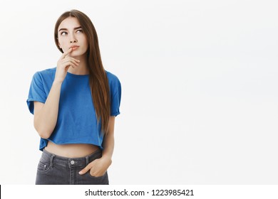 Creative smart and thoughtful attractive european woman in trendy blue cropped top making hmm gesture with finger on lower lip raising eyebrow and gazing up, thinking making choice in mind