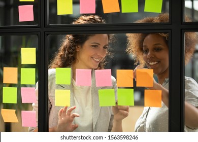 Creative smiling caucasian female team leader working together with african american millennial project manager employee intern, putting paper stickers on window glass kanban agile board at office.