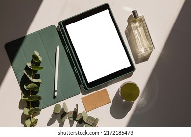 Creative space with tablet and blank screen, flowers, business card and perfume. Woman designer workspace mockup, top view. Natural light with shadows.