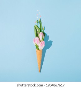 Creative spring flat lay with ice cream cone, green leaves, pink hearts and pastel tulips on blue background. Minimal concept.