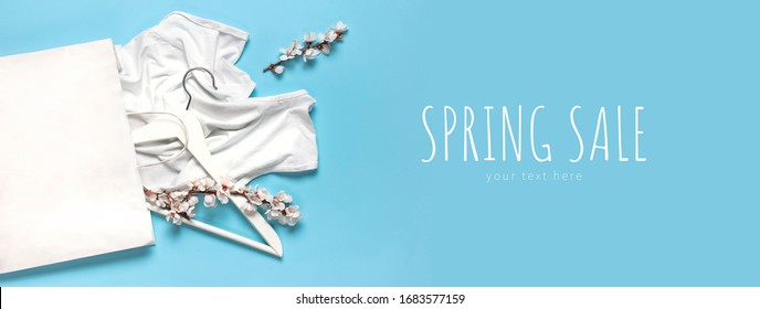 Creative Spring Sale Concept. White Wooden Hangers With Spring Flowers White Shirt Paper Bag On Blue Background Top View Flat Lay. Fashion Spring Discounts Shopping Sale Store Promo Design