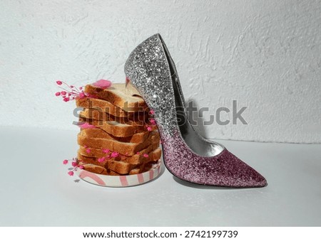 Creative still life composition featuring a pink glitter sigh shoe placed next to stacked bread slices decorated with small pink flowers. Minimalist white background and strong shadows create a playful surreal fashion concept combining food and style aestetics.