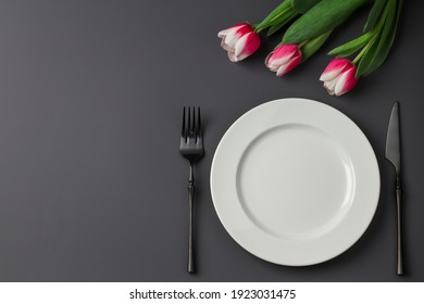 Creative table setting with white plate, black fork and knife, tulips on a dark background. Valentine's Day, Wedding Day, Birthday, Women's Day and Mother's Day. Flat lay.Copy space.