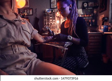Creative tattoo master with dreadlocks is creating beautiful tattoo on woman's hand at tattoo salon.