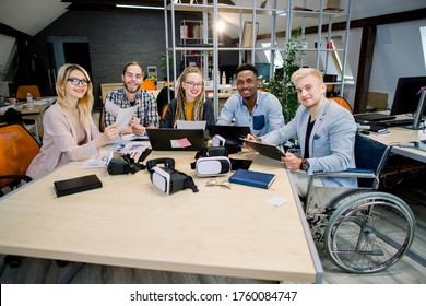 Creative Team Of Diverse Business People Sitting At Office Table During Meeting, Looking At Camera With Smile, Passionate About Work. A Handicapped Man In A Wheelchair, Chief With His Team