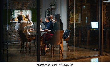 Creative Team Is Having A Conference Video Call In Meeting Room Behind Glass Walls In An Office. Colleagues Sit Behind Table And Discuss Business Opportunities With Colleague On Big TV Screen.