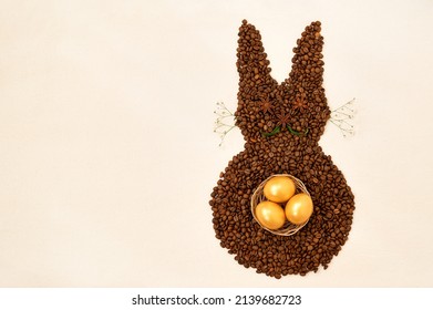 Creative Top View Flat Lay Of Easter Bunny Shape Made Of Coffee Roasted Beans And Gold Painted Eggs On Rustic Table Cloth. Rabbit Is Decorated With Anise Star Seeds, Baby's Breath Flowers. Copy Space.