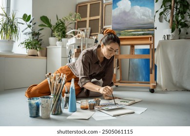 Creative woman artist freelance illustrator painting with watercolors in sketchbooks in craft workshop near easel. Concentrated inspired female painter drawing sitting on floor in modern art studio. - Powered by Shutterstock