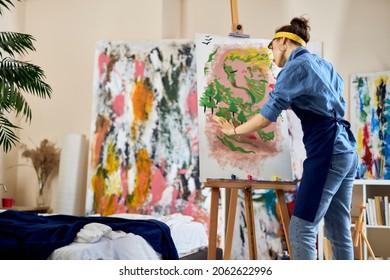 Creative woman, female artist in apron looking inspired while creating her own abstract painting at home studio workshop