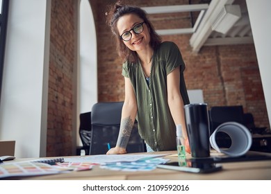 Creative woman, interior designer or architect in casual wear with messy hairdo smiling at camera while working on a blueprint for new project, standing in her office
