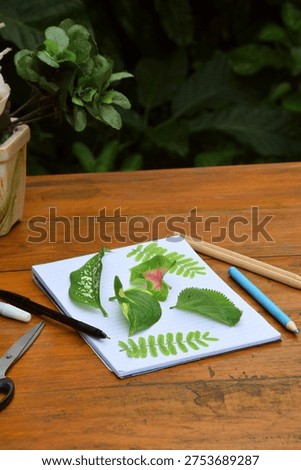 A creative workspace featuring various green leaves on a notebook with pencils, pens, and scissors on a rustic wooden table.
