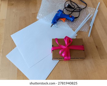A creative workspace featuring a wrapped gift with a pink bow, alongside a hot glue gun, glue sticks, and protective bubble wrap on a wooden desk.