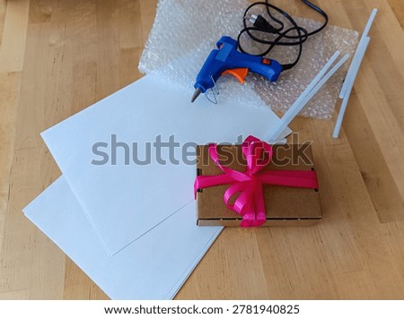 A creative workspace featuring a wrapped gift with a pink bow, alongside a hot glue gun, glue sticks, and protective bubble wrap on a wooden desk.
