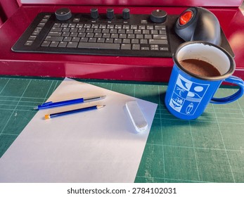 A creative workspace setup featuring a green cutting mat with a blank white sheet of paper, two pens, and a clean white eraser arranged neatly on the surface. A blue mug with a warm drink adds a cozy touch, while a keyboard with control knobs and a large trackball mouse sit in the background, blending digital tools with traditional stationery