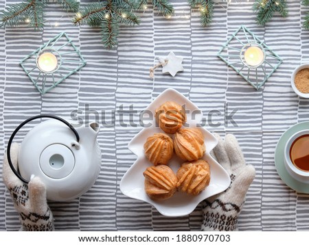 Creative Xmas table setup for tea with cakes. Creative flat lay with yummy eclairs or brewing cakes. Christmas tree shape plate with cakes. Fir twigs with light garland on stripy textile tablecloth.