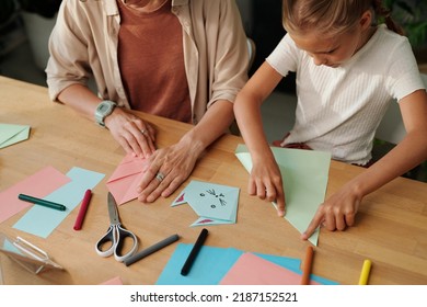 Creative youthful girl and her mother folding multi-color paper while sitting by table and making origami at leisure at home