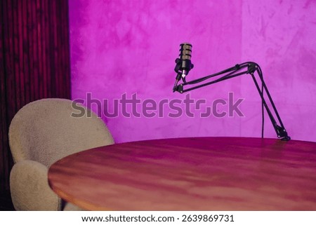 In a creatively designed broadcast space, a microphone is mounted on an adjustable arm above a wooden table, beside a cozy chair. The purple backdrop adds an inviting atmosphere for streaming.