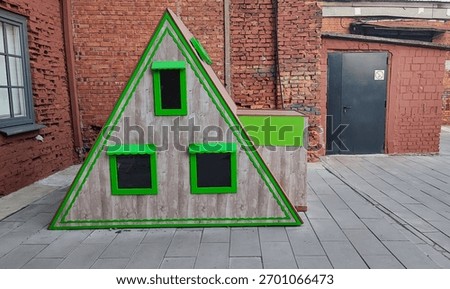 A creatively designed triangular playhouse with bright green accents is situated on a paved area beside a red brick wall.
