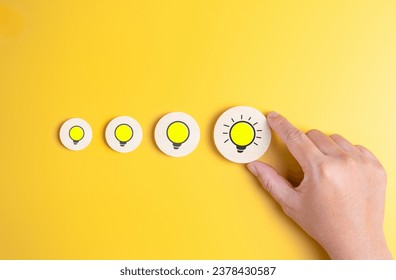Creativity ideas for solving problem concept. Hand pick the biggest wooden label with the brightest light bulb among the others on a yellow background. Creative process to finding problem solution.
