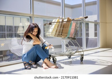 Credit Card Debt Problem. Sad Asian Woman Sitting On Floor In Mall With Shopping Trolley Cart, Shopping Bags. She Is Holding Mock Up Credit Cards Looking Upset As No Money Left And Checking In A Bill.