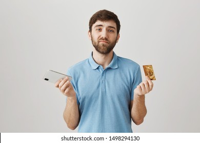 Credit Card Stopped Working. Indoor Shot Of Upset Disappointed Ordinary Caucasian Guy With Beard Holding Banking Card And Smartphone, Being Sad. Man Has Low Balance And Can Not Afford Anything