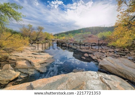 Creek at Tucson, Arizona with a view of mountain against the cloudy sky reflecting on the water. There are large rocks and trees surrounding the water.