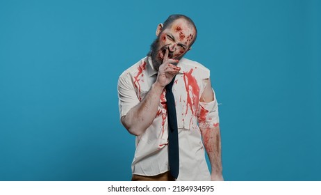 Creepy Dead Walking Corpse Making Shush Secrecy Hand Gesture On Blue Background. Dangerous Walking Dead Corpse With Deep And Bloody Wounds Being Confidential While Smirking At Camera.