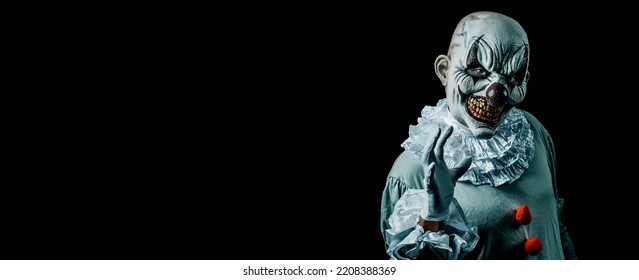 a creepy evil clown, in a gray costume with a white ruff around his neck, against a black background with some blank space on the left, in a panoramic format to use as web banner or header - Shutterstock ID 2208388369