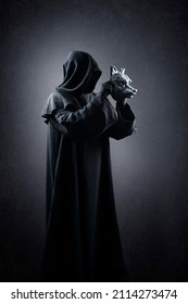 Creepy figure in hooded cloak with wolf mask in hands over dark misty background