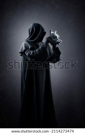 Creepy figure in hooded cloak with wolf mask in hands over dark misty background