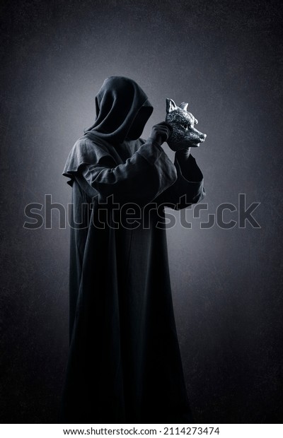 Creepy figure in hooded cloak with wolf mask\
in hands over dark misty\
background